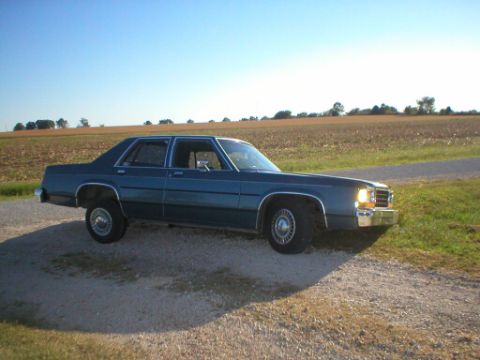 1987 Ford Crown Victoria LTD - A blue sedan is parked on a dirt road with a field behind it under a clear sky.