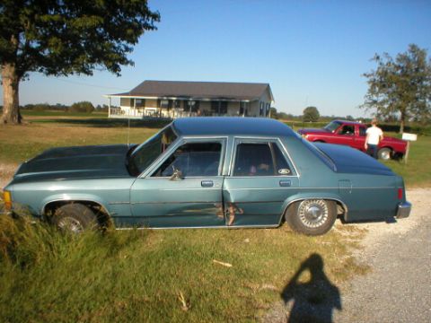 1987 Ford Crown Victoria LTD - A teal 1987 Ford Crown Victoria LTD is parked on the side of a gravel road, showing significant damage to the driver's side door.