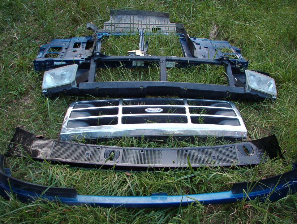 Truck front-end parts laid in grass: core support, headlights, chrome grille with Ford emblem, bumpers