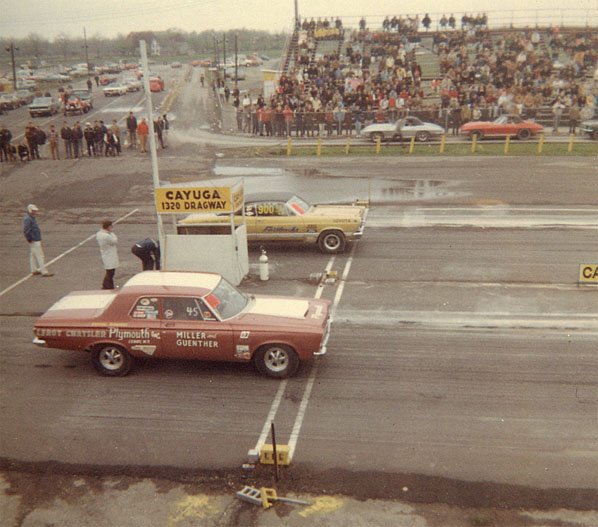 Maroon Plymouth drag car and yellow car staged at Cayuga 1320 Dragway with crowd behind.