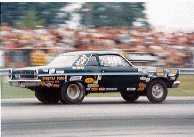 Green vintage drag racing coupe doing a wheelie on a track before a crowd