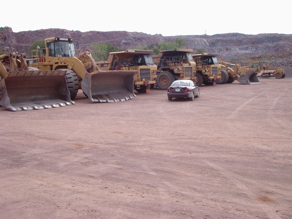 several large yellow construction trucks and a dark sedan parked on a dirt lot with a quarry in the background