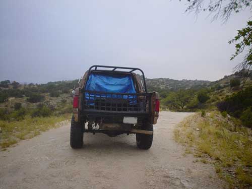 Rear view of a 1989 Ford Bronco on a dirt road with a blue tarp in the bed
