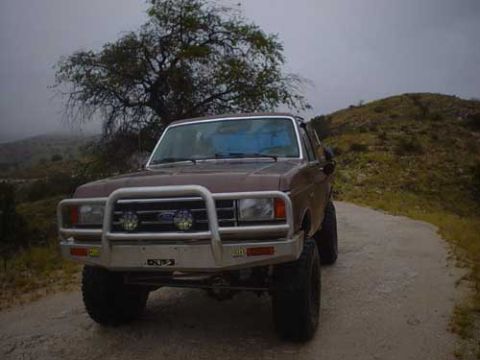 1989 Ford Bronco - This brown Ford truck, likely from the late 1980s, is parked on a dirt road with a hilly, overcast background.