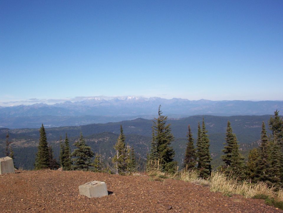 View from a gravel mountain summit over forested ridges toward distant snowy peaks; no vehicle shown