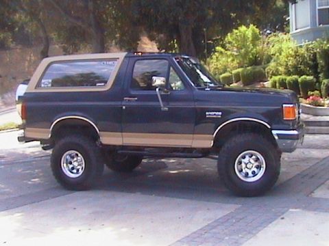 1991 Ford Bronco - This Ford Bronco is parked on a paved surface with trees and a house in the background.