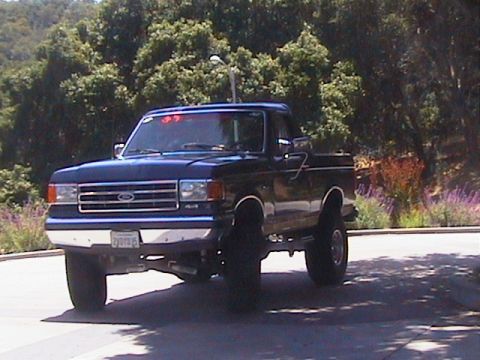1991 Ford Bronco - Three-quarter front view of a dark blue lifted Ford Bronco with the hardtop removed, parked on a paved road with wooded hillside and purple…