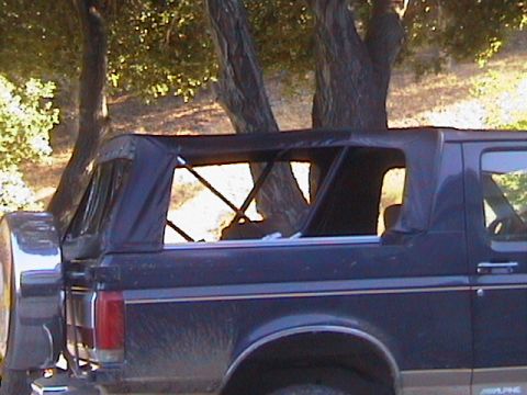 1991 Ford Bronco - The side profile of a Ford Bronco shows its soft top enclosure and spare tire, set against a backdrop of trees.