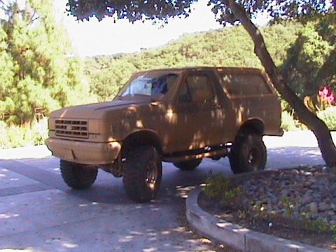 1991 Ford Bronco - This lifted 1987 Ford Bronco sits parked outdoors under a tree, its tan paint faded and worn.