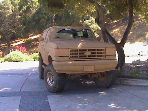 1991 Ford Bronco - Lifted Ford Bronco parked in a driveway completely coated in dried tan mud from bumper to roof after an off-road run, viewed from the front…