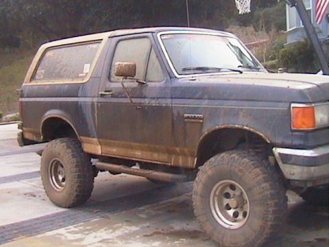 1991 Ford Bronco - Close side view of a lifted blue and tan Ford Bronco splattered with drying mud along the doors, rocker panels and hardtop, parked on a wet…