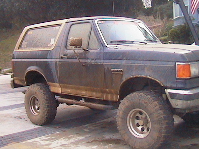 Mud-splattered lifted blue Ford Bronco on white-spoke wheels, side view