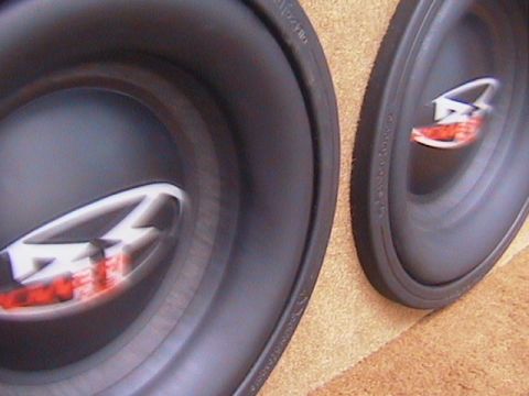 1991 Ford Bronco - Close-up of a pair of black car-audio subwoofers lying face-up on tan carpet, their dust caps stamped with a chrome and red logo. The rubbe…