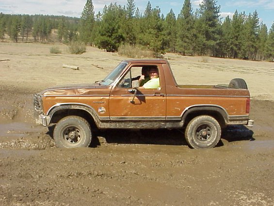 Brown 1981 Ford Bronco with top removed, side view, driving through deep mud with pine trees behind