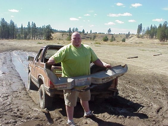 Man in green shirt holds a steel bumper while standing by a mud-covered small pickup in an open field