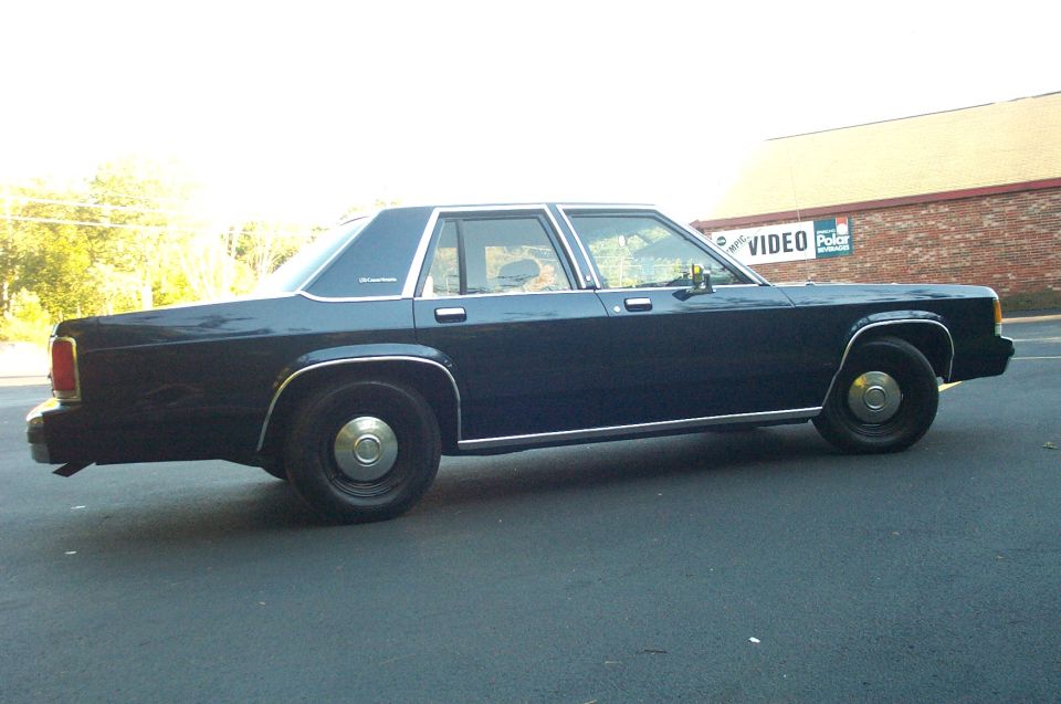 Dark blue sedan parked on asphalt, side view. Chrome trim along the bottom and windows. "Crown Victoria" badge visible on rear fender