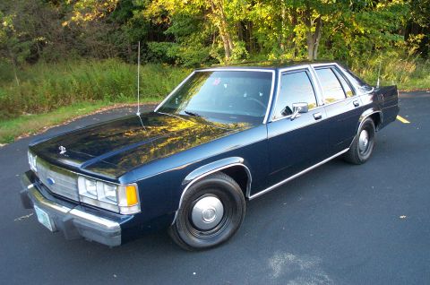 1991 Ford LTD Crown Victoria - A dark blue sedan with chrome accents is parked on an asphalt surface, with trees and foliage in the background.