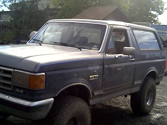 Lifted two-tone gray and blue Ford Bronco SUV with large off-road tires, front three-quarter view on gravel