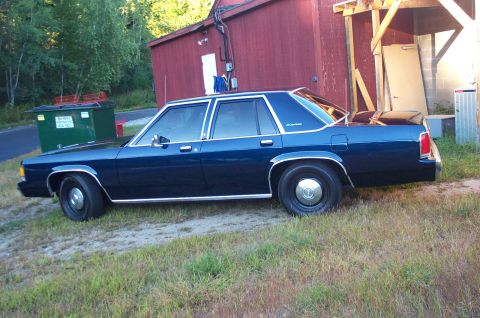 1991 Ford LTD Crown Victoria - A dark blue sedan, likely a 1990 model, is parked outdoors next to a red building.