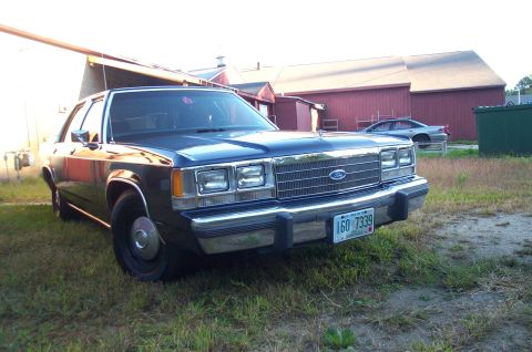 1991 Ford LTD Crown Victoria - A dark blue Ford Crown Victoria is parked in front of a red barn, with another car visible in the background.