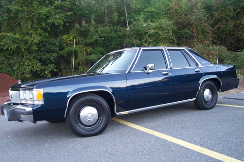1991 Ford LTD Crown Victoria - A dark blue Ford LTD Crown Victoria four-door sedan shown in side/3-quarter front view, parked on asphalt with yellow parking lines and gre…