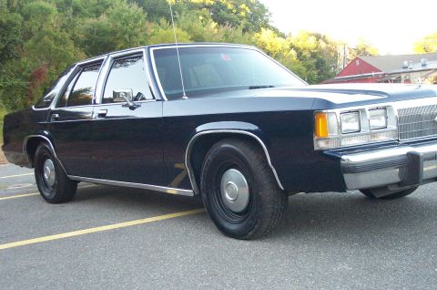 1991 Ford LTD Crown Victoria - A dark blue sedan is shown from the front quarter angle, parked in a lot with trees in the background.