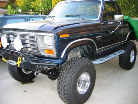 1985 Ford Bronco - This lifted Ford Bronco features prominent off-road lights and protective rock sliders, with "SUPERMOTORS" visible on the lower right.
