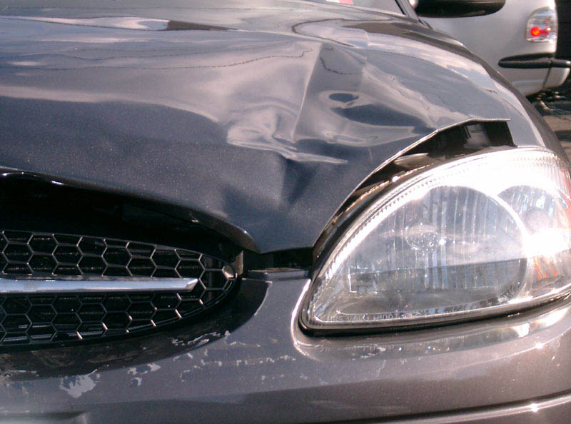 Dark gray sedan with crumpled, dented hood above passenger headlight and grille after front-end damage