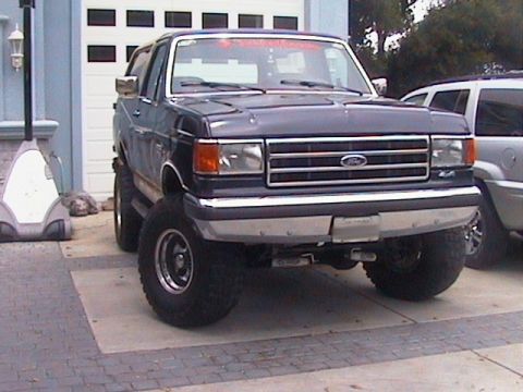 1991 Ford Bronco - This lifted 1991 Ford Bronco features a dark blue exterior and prominent large tires, seen from the front.
