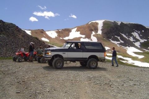 1993 Ford Bronco - A two-tone Ford Bronco is parked on a rocky mountain road with ATVs nearby.