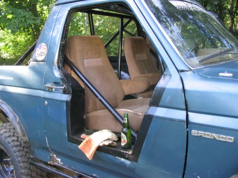 1986 Ford Bronco - The interior of a blue SUV features a roll cage and tan seats, with a bottle of beer and gloves visible.