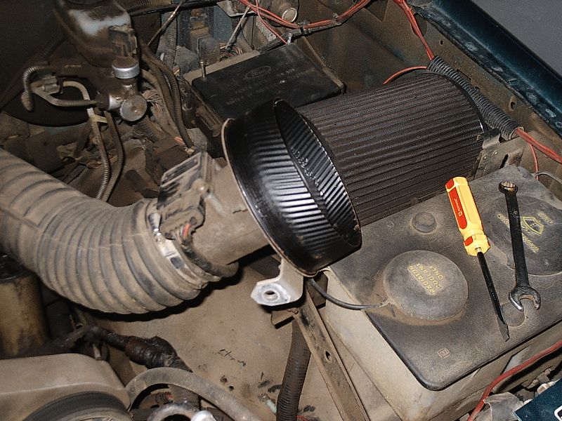 Aftermarket cone air filter and intake tube in a dusty engine bay with tools on the battery.