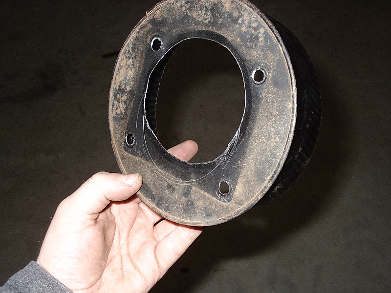 Hand holding a dirty round metal flanged part with four bolt holes and oval center