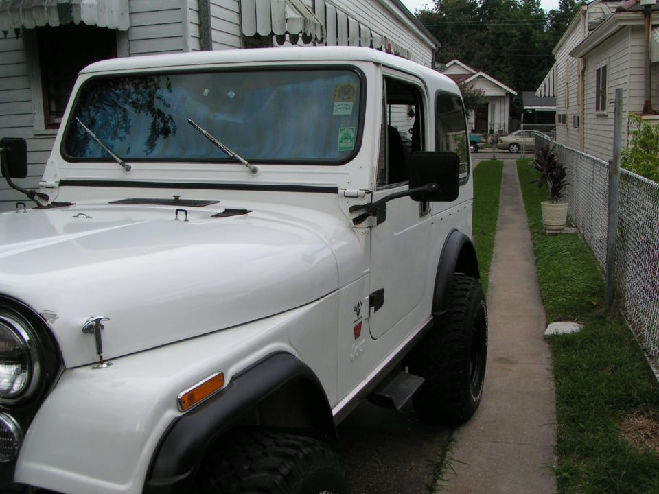 White Jeep CJ7 hardtop parked on driveway between houses, front 3/4 view