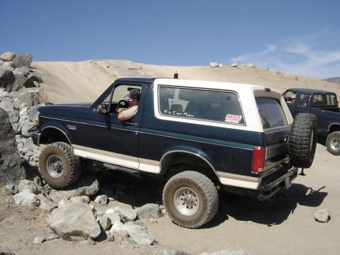 1988 Ford Bronco - A Ford Bronco is shown tackling a rocky trail with a pickup truck in the background under a clear blue sky.