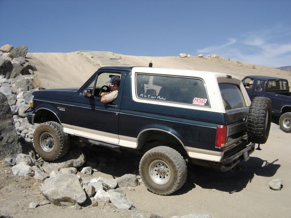 Dark blue and white Ford Bronco navigating rocky terrain. Another dark pickup truck is visible behind it