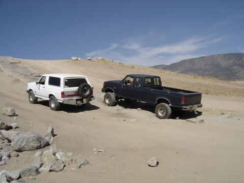 1988 Ford Bronco - Two off-road vehicles, a white SUV and a dark blue pickup truck, navigate a dusty, mountainous terrain.
