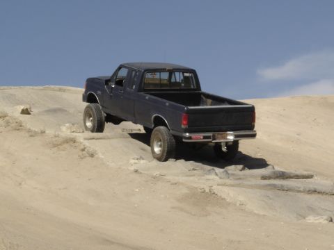 1988 Ford Bronco - A dark blue pickup truck navigates a rocky dirt incline under a clear blue sky.