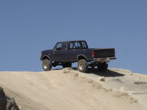 1988 Ford Bronco - A lifted Ford pickup truck navigates a rocky, off-road terrain under a clear blue sky.