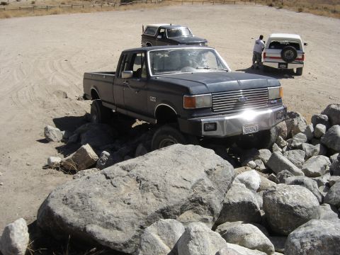 1988 Ford Bronco - A dark blue Ford pickup truck is shown off-roading over rocks, with two Ford Broncos in the background.