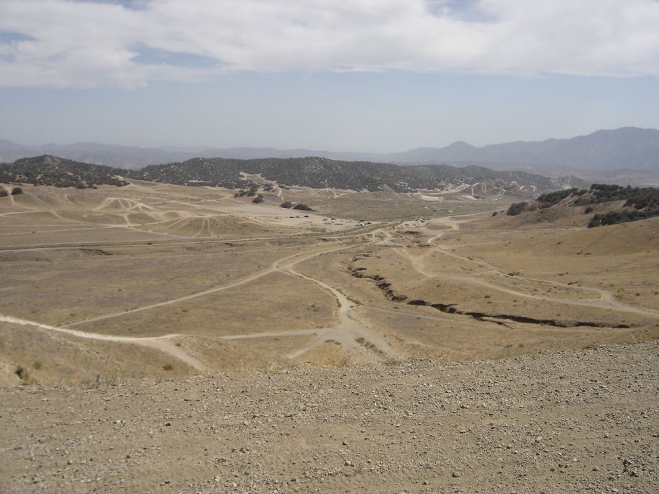 Dirt roads wind through a dry, hilly landscape under a hazy sky; two vehicles are visible in the distance