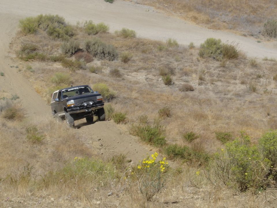 Lifted tan Ford Bronco climbing a dirt hill trail on big mud tires in dry desert brush