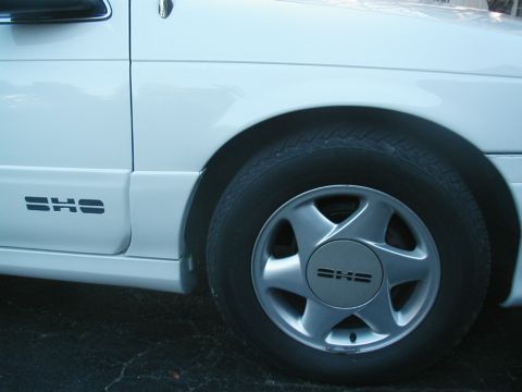 1993 Ford Taurus - The side profile of a white 1993 Ford Taurus SHO is visible, its distinctive wheel and door emblem.