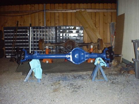 1988 Ford Bronco - Two vehicle axles are visible, one freshly painted blue and the other rusty, set up in a garage-like setting.