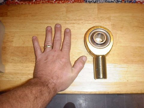1988 Ford Bronco - A large gold cadmium-plated heim (rod end) joint with a coarse-threaded male shank lying on a butcher-block table, with an open hand placed…