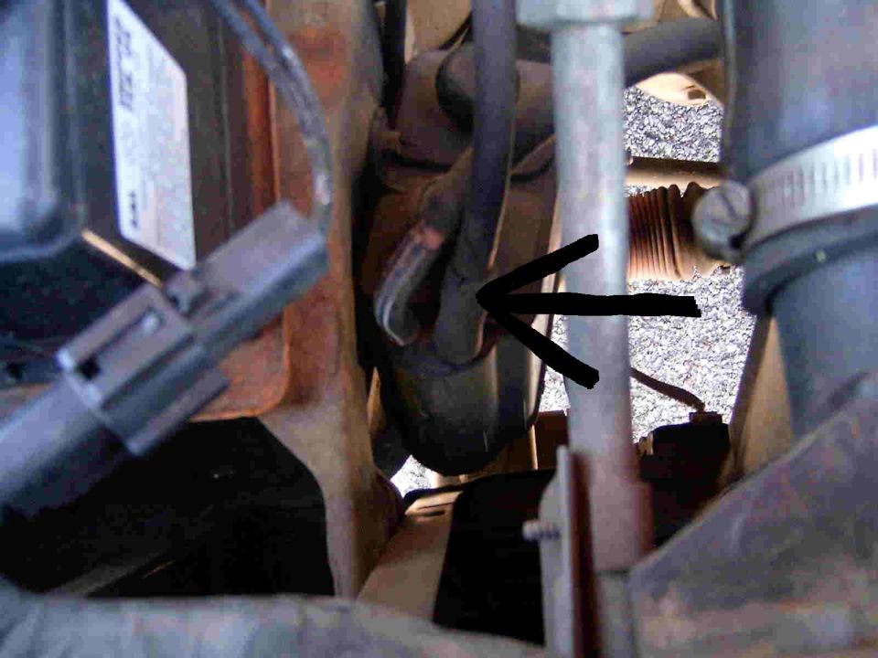 Engine bay close-up with black arrow pointing to a component among hoses and wiring