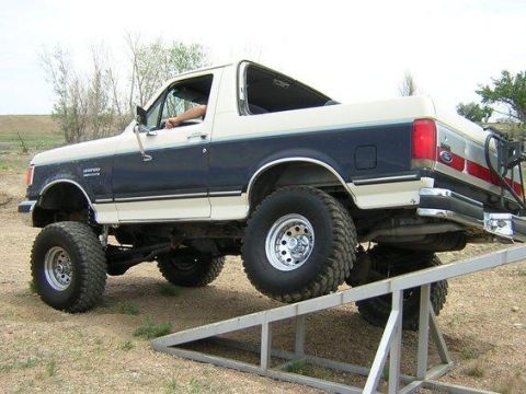1988 Ford Bronco - A lifted two-tone dark blue and white 1988-era Ford Bronco full-size SUV shown from the rear three-quarter angle with its front wheels driv…