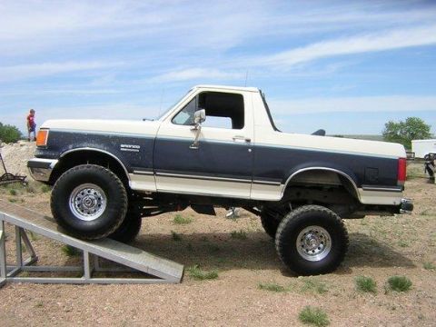 1988 Ford Bronco - Side profile of a two-tone dark blue and white Ford Bronco full-size SUV with a lifted suspension and large off-road tires, parked on dirt…