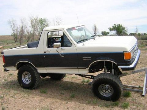 1988 Ford Bronco - A two-tone white over dark blue 1988 Ford Bronco shown in side profile with a suspension lift, oversized mud-terrain tires, and chrome slot…