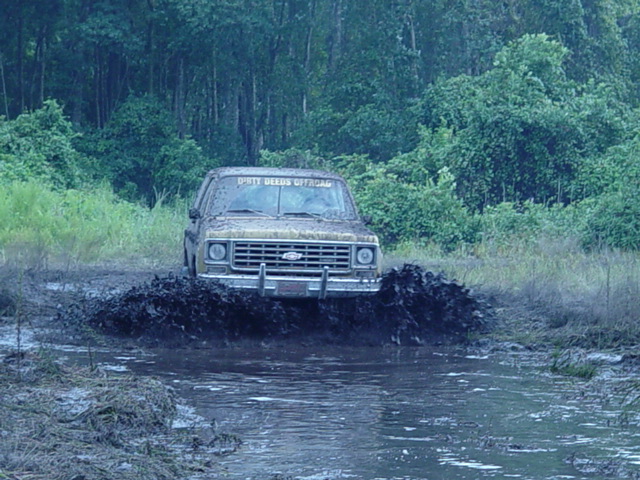 Tan pickup truck with "DIRTY DEEDS OFFROAD" on windshield driving through mud puddle
